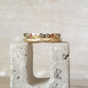 May include: Two gold rings stacked on a concrete block. The top ring is smooth and set with small, colorful gemstones. The bottom ring has a textured surface. The gemstones are red, blue, and yellow.