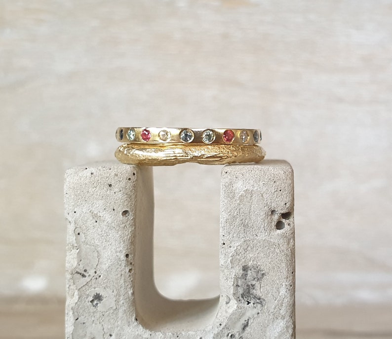May include: Two gold rings stacked on a concrete block. The top ring is smooth and set with small, colorful gemstones. The bottom ring has a textured surface. The gemstones are red, blue, and yellow.