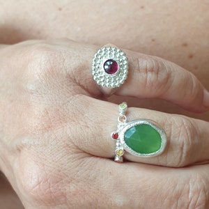 May include: Two silver rings with colorful gemstones. The top ring features a red gemstone surrounded by a circle of small silver beads. The bottom ring has a green gemstone with a silver setting and a row of colorful gemstones on the band.