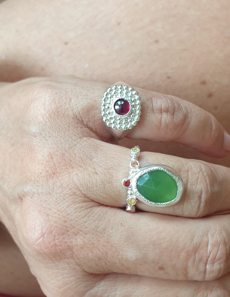 May include: Two silver rings with colorful gemstones. The top ring features a red gemstone surrounded by a circle of small silver beads. The bottom ring has a green gemstone with a silver setting and a row of colorful gemstones on the band.