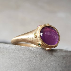 K&ouml;nnte beinhalten: Ein goldener Ring mit einem gro&szlig;en violetten Edelstein, umgeben von kleineren gr&uuml;nen, gelben und rosa Edelsteinen.