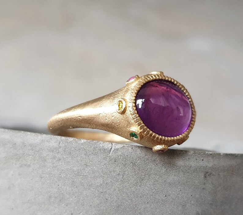 K&ouml;nnte beinhalten: Ein goldener Ring mit einem gro&szlig;en violetten Edelstein, umgeben von kleineren gr&uuml;nen, gelben und rosa Edelsteinen.