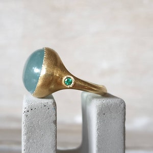 May include: A gold ring with a large, oval-shaped aquamarine gemstone and a small, green emerald accent stone.