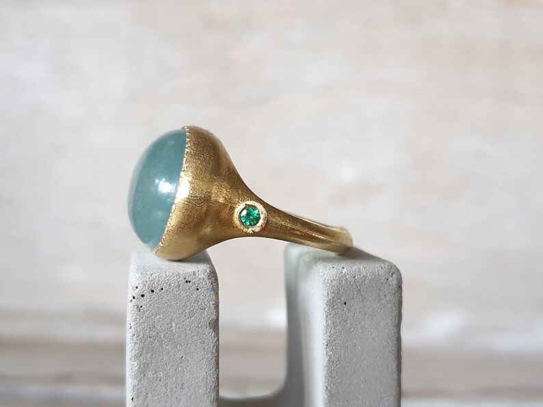 May include: A gold ring with a large, oval-shaped aquamarine gemstone and a small, green emerald accent stone.