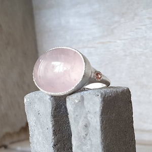 Puede incluir: Un anillo de plata con una gran piedra de cuarzo rosa ovalada. El anillo tiene un borde texturizado y una pequeña gema redonda de color rosa anaranjado en la banda. El anillo se muestra sobre una superficie gris texturizada.