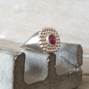 May include: A silver ring with a red gemstone set in a textured, circular bezel. The ring is on a gray concrete surface.