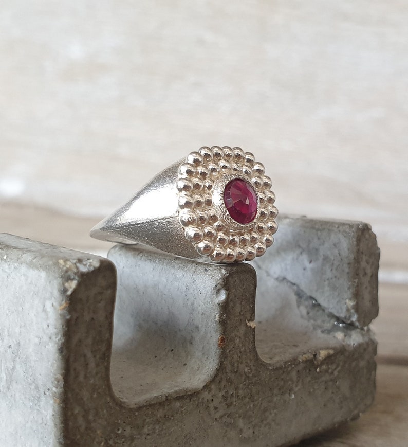 May include: A silver ring with a red gemstone set in a textured, circular bezel. The ring is on a gray concrete surface.