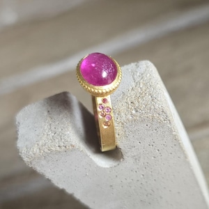 Op de afbeelding: Een gouden ring met een grote, ovale, magenta edelsteen. De ring heeft een getextureerde gouden band en is versierd met kleinere roze edelstenen. De ring staat tegen een neutrale achtergrond.
