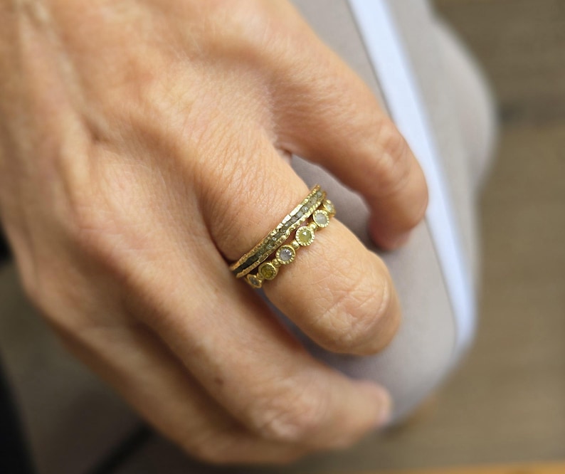 Pu&ograve; includere: Tre anelli d'oro con piccoli diamanti incastonati in fila. Gli anelli sono impilati su un dito.