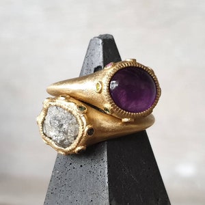 K&ouml;nnte beinhalten: Zwei goldene Ringe mit Edelsteinen. Der obere Ring hat einen gro&szlig;en, runden, violetten Amethyststein, der von kleineren, bunten Steinen umgeben ist. Der untere Ring hat einen gro&szlig;en, runden, klaren, facettierten Stein, der von kleineren, bunten Steinen umgeben ist.