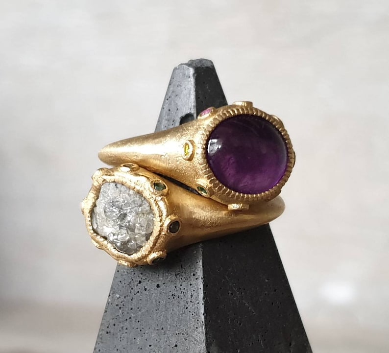 K&ouml;nnte beinhalten: Zwei goldene Ringe mit Edelsteinen. Der obere Ring hat einen gro&szlig;en, runden, violetten Amethyststein, der von kleineren, bunten Steinen umgeben ist. Der untere Ring hat einen gro&szlig;en, runden, klaren, facettierten Stein, der von kleineren, bunten Steinen umgeben ist.