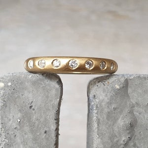 Peut inclure: Une bague en or avec une finition mate, sertie de plusieurs petites pierres pr&eacute;cieuses rondes et claires. La bague repose sur une surface grise et textur&eacute;e. Les pierres sont espac&eacute;es uniform&eacute;ment.