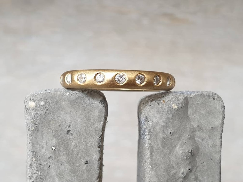Peut inclure: Une bague en or avec une finition mate, sertie de plusieurs petites pierres pr&eacute;cieuses rondes et claires. La bague repose sur une surface grise et textur&eacute;e. Les pierres sont espac&eacute;es uniform&eacute;ment.
