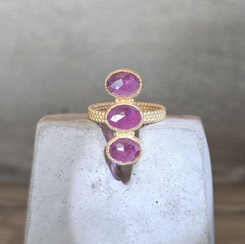 May include: A gold ring featuring three vertically aligned, oval-shaped, faceted pink gemstones. The band has a textured, beaded surface, and each stone is set within a delicate gold bezel. The design creates a linear arrangement of stones.