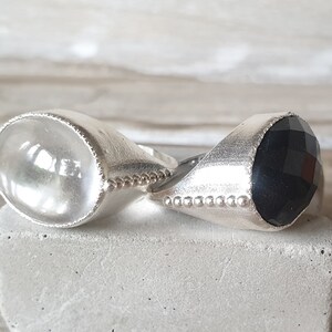 May include: Two silver rings with gemstone settings. The ring on the left has a clear, faceted gemstone. The ring on the right has a black, faceted gemstone.
