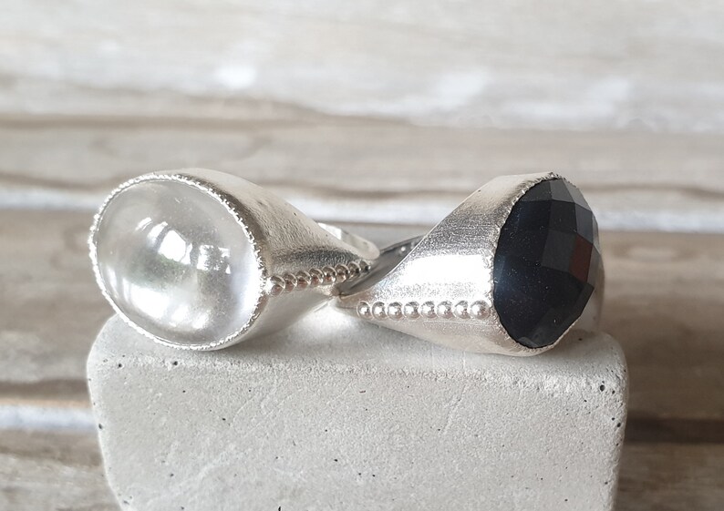 May include: Two silver rings with gemstone settings. The ring on the left has a clear, faceted gemstone. The ring on the right has a black, faceted gemstone.