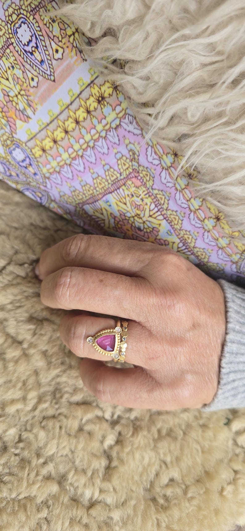 May include: A gold ring with a triangular, magenta gemstone and small white stones. The ring is on a finger, with a second gold band visible. The background includes a patterned fabric and a fluffy, light-colored surface.