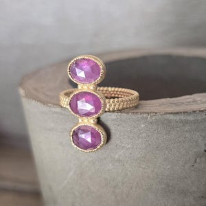 May include: A gold ring featuring a textured, beaded band and three vertically aligned, oval-shaped, faceted magenta-pink gemstones. The stones are set in gold bezels with a fine milgrain edge, creating a linear design across the finger.