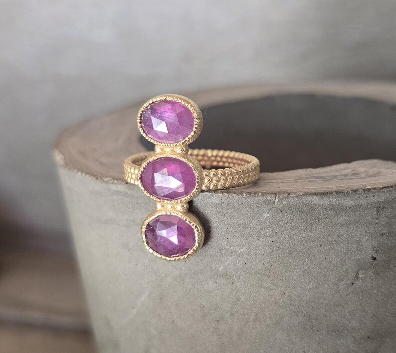 May include: A gold ring featuring a textured, beaded band and three vertically aligned, oval-shaped, faceted magenta-pink gemstones. The stones are set in gold bezels with a fine milgrain edge, creating a linear design across the finger.