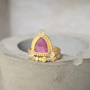 May include: A gold ring set featuring a triangular pink gemstone and small diamonds. The main ring has a beaded edge and a triangular pink stone. A smaller ring with a row of gold circles and diamonds sits below.