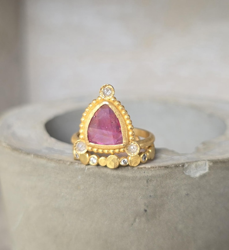 May include: A gold ring set featuring a triangular pink gemstone and small diamonds. The main ring has a beaded edge and a triangular pink stone. A smaller ring with a row of gold circles and diamonds sits below.