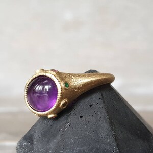 K&ouml;nnte beinhalten: Ein goldener Ring mit einem gro&szlig;en, runden, violetten Amethyst-Edelstein, der von kleineren gr&uuml;nen und roten Edelsteinen umgeben ist.