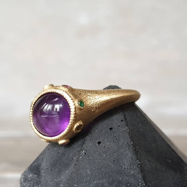 K&ouml;nnte beinhalten: Ein goldener Ring mit einem gro&szlig;en, runden, violetten Amethyst-Edelstein, der von kleineren gr&uuml;nen und roten Edelsteinen umgeben ist.