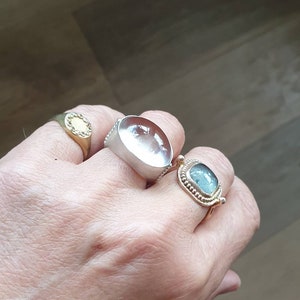 May include: A hand wearing four rings. One is gold with a clear stone, another is silver with a large, clear, oval stone, and a third is gold with a blue-green stone. The fourth ring is gold with a small, clear stone.