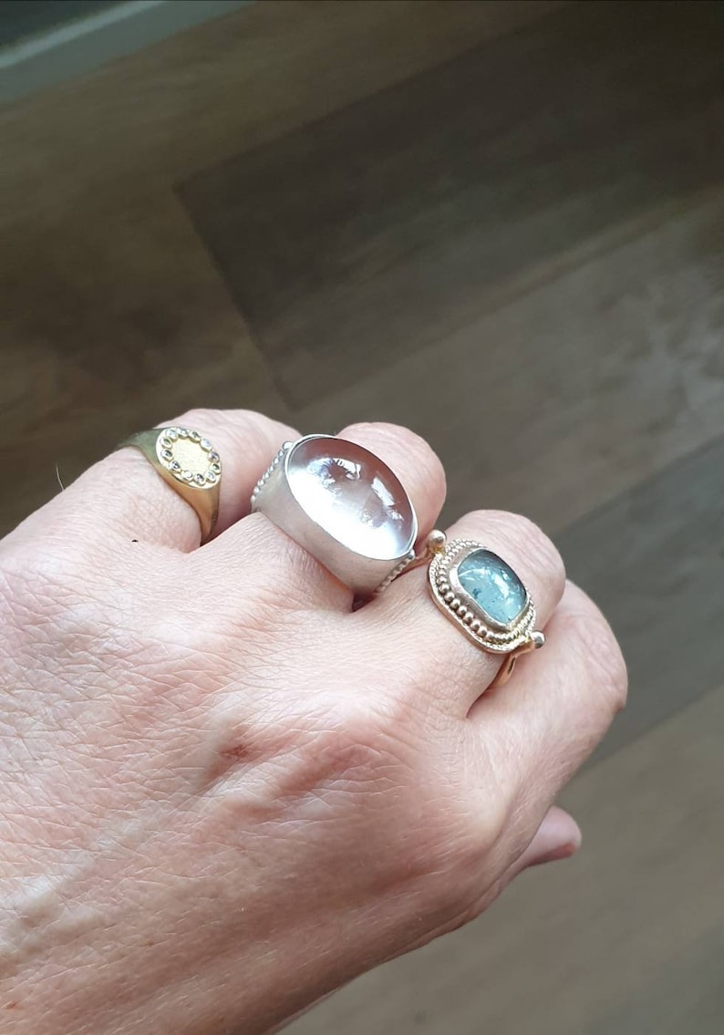 May include: A hand wearing four rings. One is gold with a clear stone, another is silver with a large, clear, oval stone, and a third is gold with a blue-green stone. The fourth ring is gold with a small, clear stone.