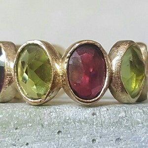 May include: A gold ring with alternating green and red oval-shaped gemstones. The ring has a textured band.