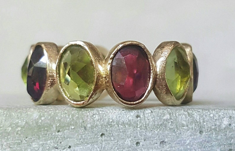 May include: A gold ring with alternating green and red oval-shaped gemstones. The ring has a textured band.