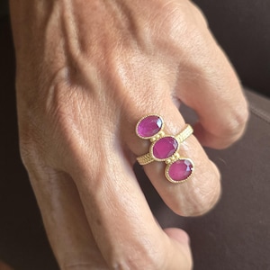 May include: A gold ring featuring three oval-shaped, vibrant pink gemstones arranged in a vertical row. The band has a textured, beaded surface, and the stones are set in individual gold bezels with small decorative gold spheres between them.
