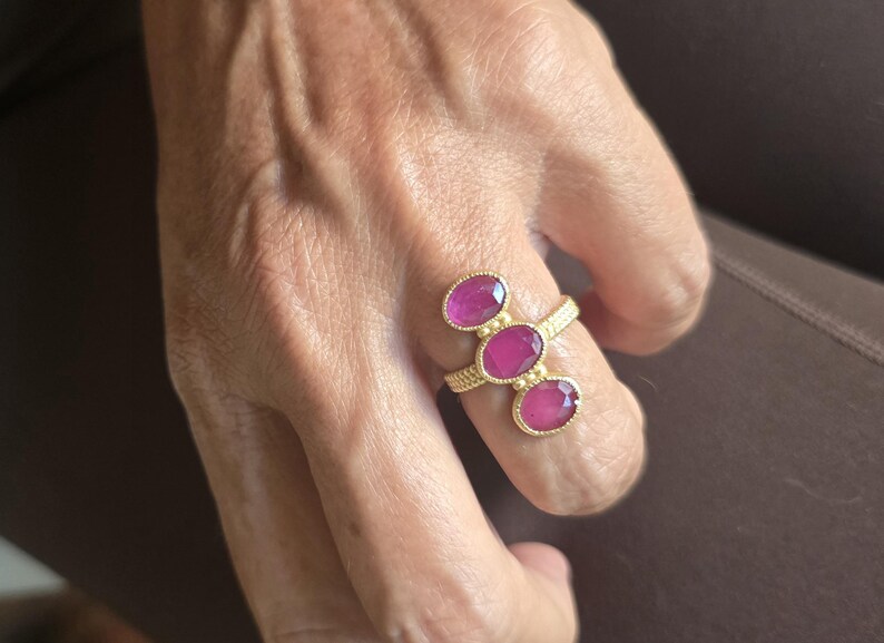 May include: A gold ring featuring three oval-shaped, vibrant pink gemstones arranged in a vertical row. The band has a textured, beaded surface, and the stones are set in individual gold bezels with small decorative gold spheres between them.