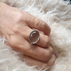 May include: A silver ring with a large, oval, clear stone. The ring is on a hand resting on a fluffy, white surface. The stone appears to be slightly translucent.