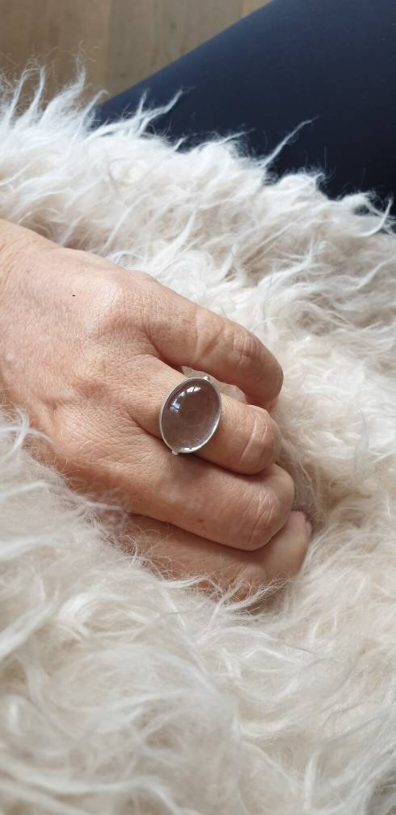 May include: A silver ring with a large, oval, clear stone. The ring is on a hand resting on a fluffy, white surface. The stone appears to be slightly translucent.