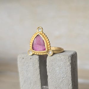 May include: A gold ring with a triangular pink gemstone and three smaller white gemstones. The ring has a textured gold band and a decorative beaded border around the pink stone. The ring is displayed on a gray, textured surface.