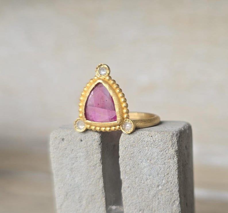 May include: A gold ring with a triangular pink gemstone and three smaller white gemstones. The ring has a textured gold band and a decorative beaded border around the pink stone. The ring is displayed on a gray, textured surface.