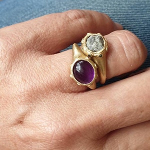 K&ouml;nnte beinhalten: Zwei goldene Ringe mit ovalen Edelsteinen. Der obere Ring hat einen grauen Stein mit rauer Textur. Der untere Ring hat einen violetten Stein mit glatter Oberfl&auml;che.
