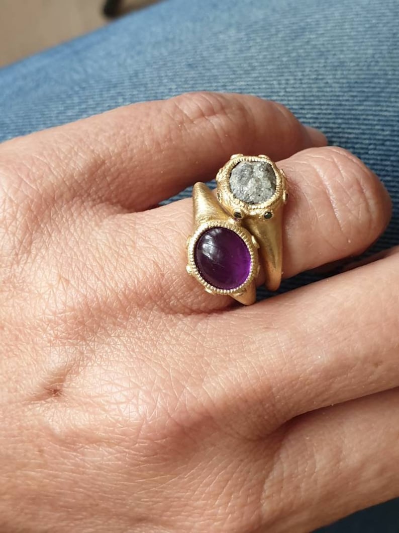 K&ouml;nnte beinhalten: Zwei goldene Ringe mit ovalen Edelsteinen. Der obere Ring hat einen grauen Stein mit rauer Textur. Der untere Ring hat einen violetten Stein mit glatter Oberfl&auml;che.