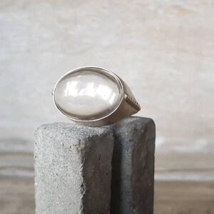 May include: A silver ring with a large, oval, pearlescent stone. The ring has a simple band with a textured design on the sides. The stone is set in a bezel setting, and the ring is displayed on a gray, textured surface.