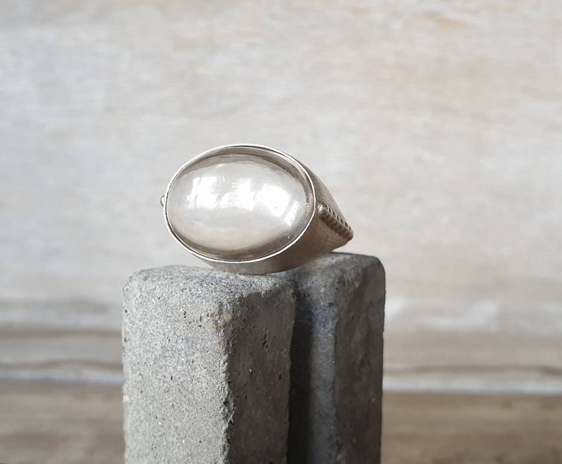 May include: A silver ring with a large, oval, pearlescent stone. The ring has a simple band with a textured design on the sides. The stone is set in a bezel setting, and the ring is displayed on a gray, textured surface.