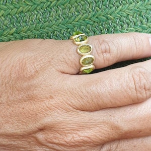 May include: A gold ring with five oval-shaped green gemstones. The ring is on a person's finger.