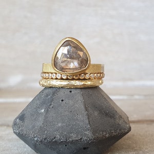 May include: A stack of gold rings with a large, triangular, light-colored gemstone. The top ring has a textured band, followed by a ring with small, clear stones, and a textured band at the bottom. The rings are displayed on a dark gray, geometric stand.