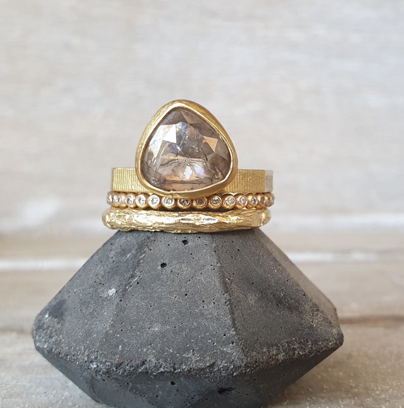 May include: A stack of gold rings with a large, triangular, light-colored gemstone. The top ring has a textured band, followed by a ring with small, clear stones, and a textured band at the bottom. The rings are displayed on a dark gray, geometric stand.
