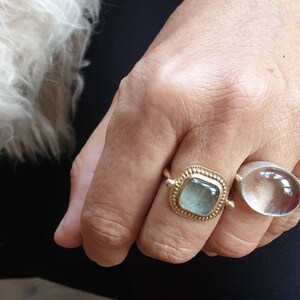 May include: Close-up of a hand wearing two rings. One ring is gold with a square, light blue stone. The other ring is silver with a large, clear, oval stone. The hand is resting on a dark blue surface.