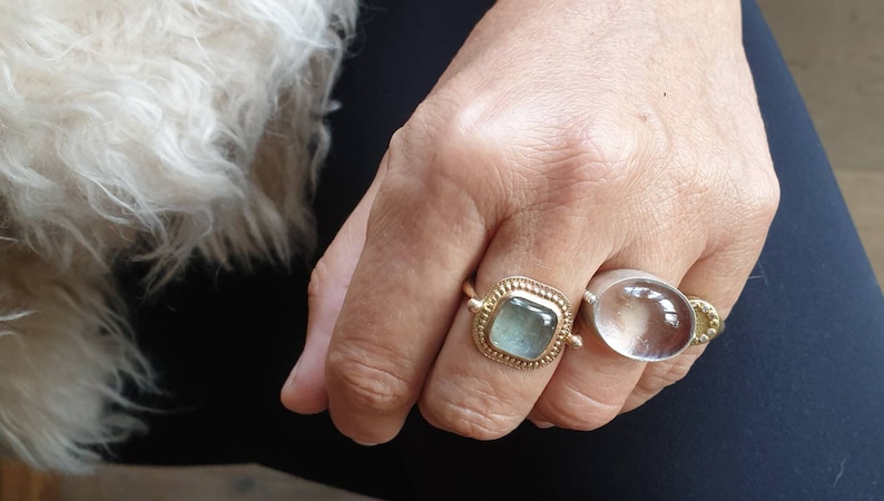 May include: Close-up of a hand wearing two rings. One ring is gold with a square, light blue stone. The other ring is silver with a large, clear, oval stone. The hand is resting on a dark blue surface.