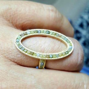 May include: A gold ring with a large oval-shaped setting. The setting is filled with small, green, and gray diamonds.