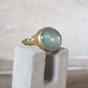 May include: A gold ring with a large, light blue aquamarine gemstone and two smaller green gemstones. The ring is on a gray stand.