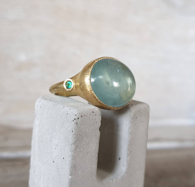 May include: A gold ring with a large, light blue aquamarine gemstone and two smaller green gemstones. The ring is on a gray stand.