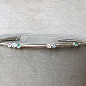 Puede incluir: Un brazalete de plata con una fila de pequeñas piedras preciosas de colores. Las piedras preciosas son rosas, azules y verdes.